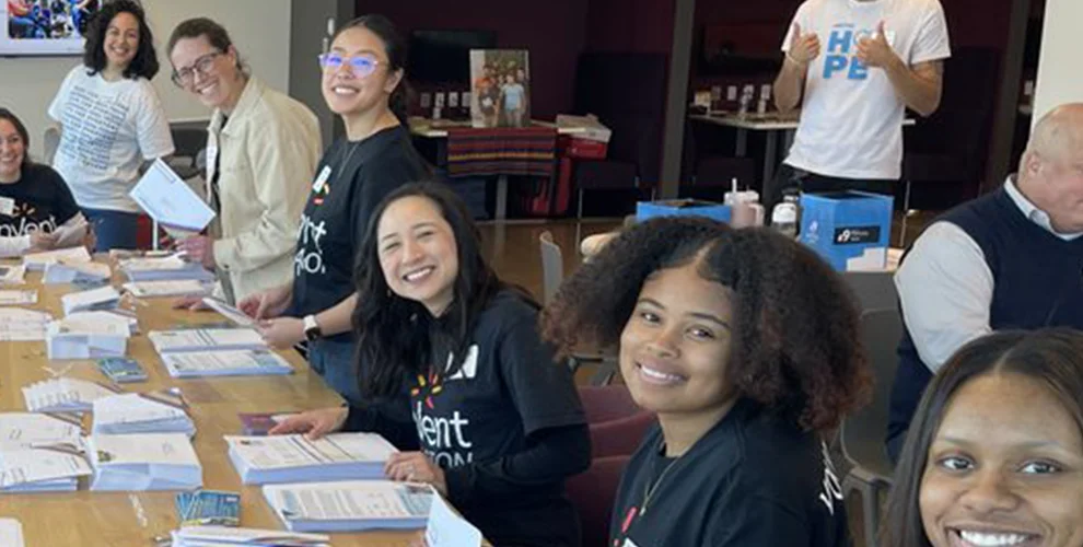 A group of people sitting around a large table working on a craft project and smiling at the camera
